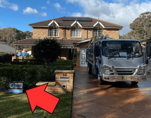 A two-story brick house with solar panels on the roof. A sign for "Penrith Solar Centre" and a work truck are in the driveway. An arrow points to the sign.