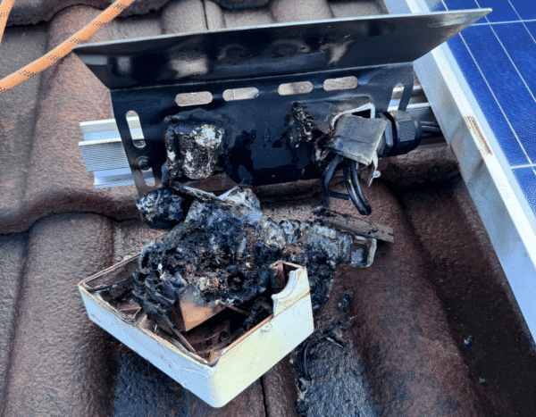 A severely burned electrical component on a roof next to a solar panel, with melted and charred wires and casing.