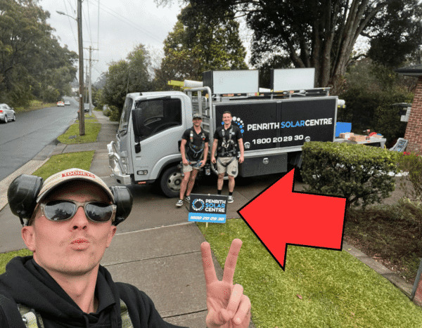 Three people in work attire with protective headgear stand near a truck bearing the logo "PENRITH SOLAR CENTRE." One person poses in the foreground and another holds a sign with the same logo.