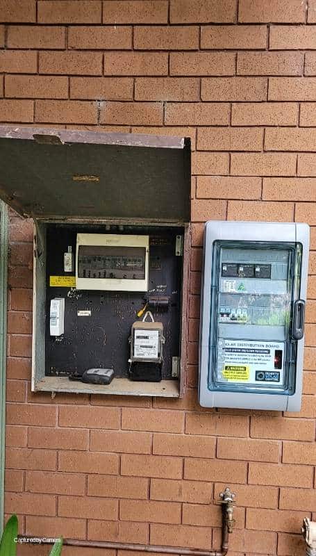 Two electrical panels are mounted on a brick wall, one open showing switches and wires, and the other closed with a transparent cover.