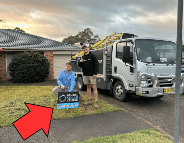 Two men in work clothes pose near a "Penrith Solar Centre" sign, with a white truck and a house in the background. The man on the left is kneeling beside the sign, while the man on the right stands.