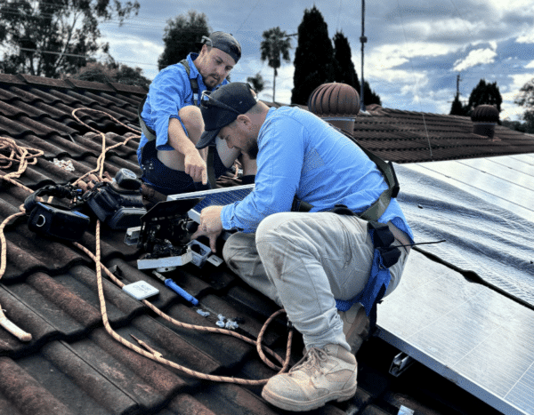 Two workers wearing blue shirts and safety gear install solar panels on a brown tiled roof under a partly cloudy sky. Various tools and cables are scattered around them.