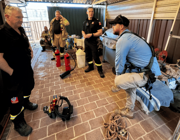 A group of firefighters in uniform stands and sits in a brick-paved area with firefighting equipment and tools around them. One person is seated on a bench wearing a harness.