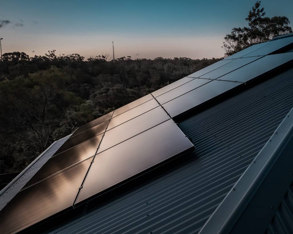 Rooftop solar panels during sunset, partially reflecting the sky, with a forested background.