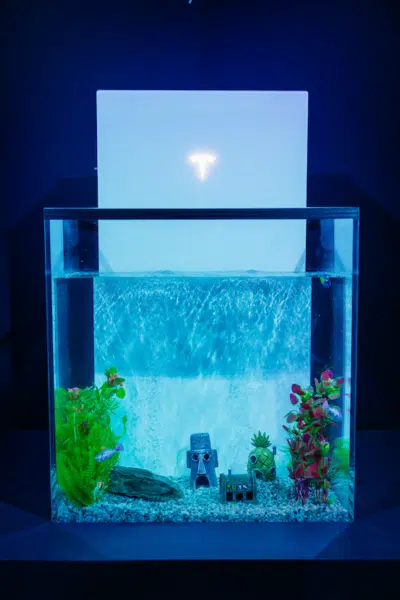 A glass aquarium with plants, decorative structures, and fish, placed in front of a glowing white Tesla logo backdrop.