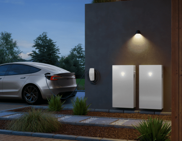 Electric car charging at a home wall charger, next to two battery storage units, outdoors during early evening.