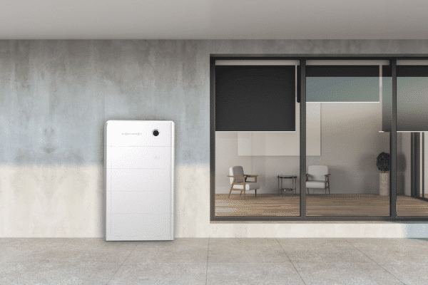 A white wall-mounted energy storage unit is next to a modern glass door leading to a room with chairs and a plant.