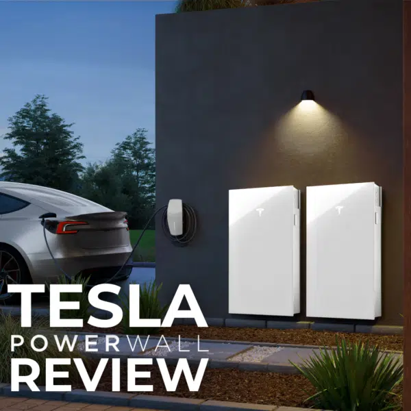 Two Tesla Powerwalls installed on an exterior wall beside a charging Tesla car. Text reads "Tesla Powerwall Review." A lit wall-mounted lamp is above the Powerwalls.