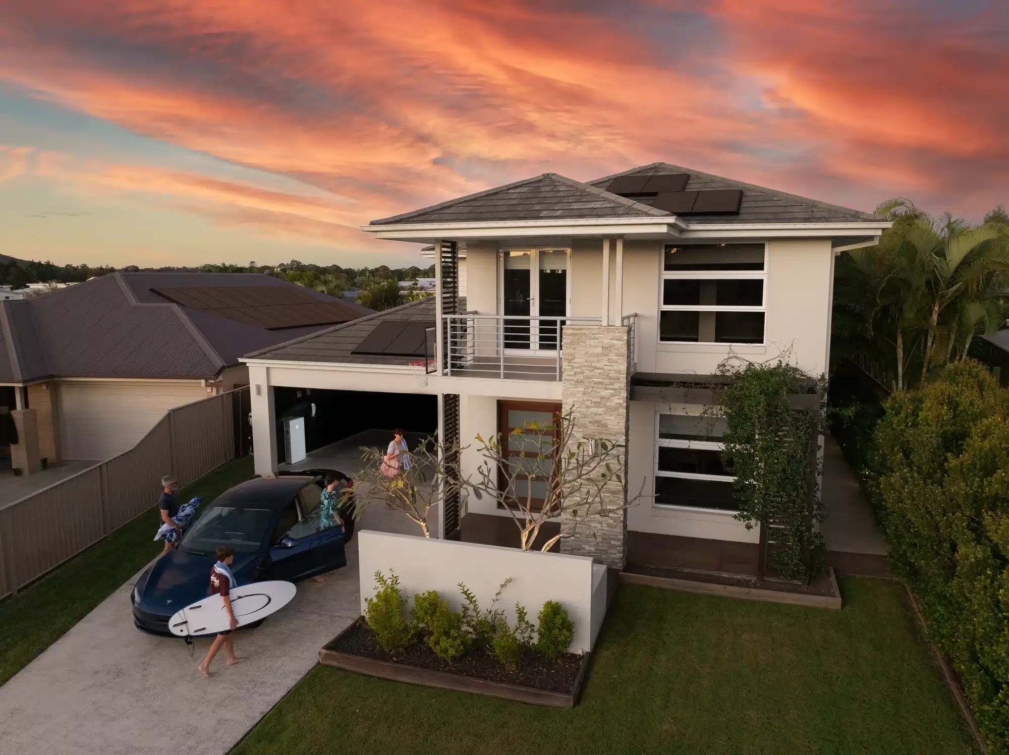 A modern two-story house with solar panels, a driveway with a parked convertible, and a person holding a surfboard. The sky is tinged with vibrant hues of orange, pink, and purple during sunset.