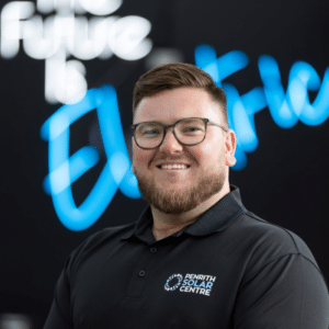 A smiling man in a black polo shirt standing in front of a blurred sign that reads "the future is electric."