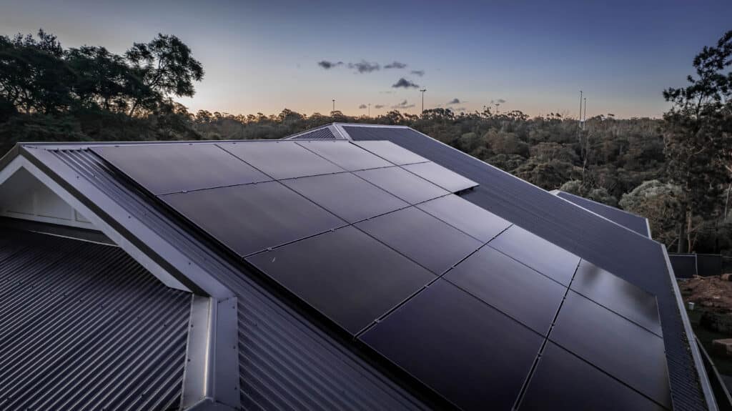 Solar panels installed on a sloped house roof at dusk, surrounded by trees and an evening sky in the background.