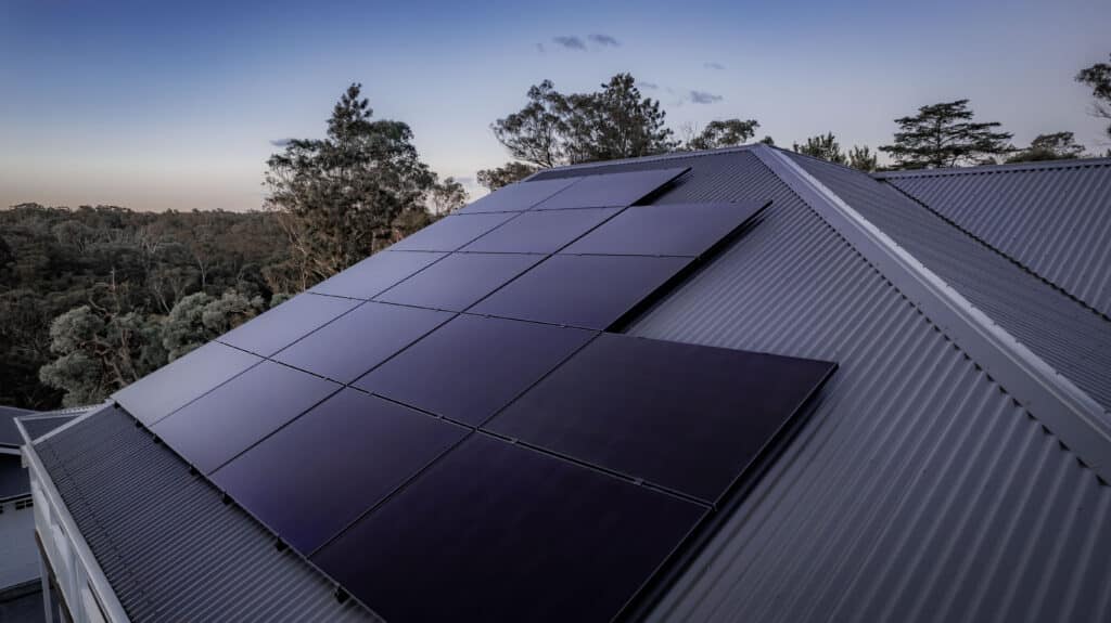 Solar panels installed on a sloped metal roof with trees and a clear sky in the background.