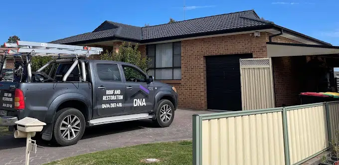 A black pickup truck with "DNA Roof and Restoration" signs is parked in the driveway of a single-story brick house with a dark tiled roof. A ladder is mounted on the truck, and a small mailbox is in the foreground.