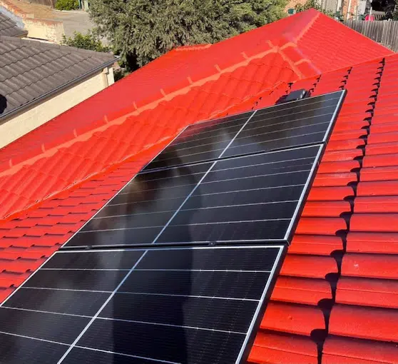 A section of a rooftop with red tiles showing an installed solar panel system under the sun. Houses and trees are visible in the background.