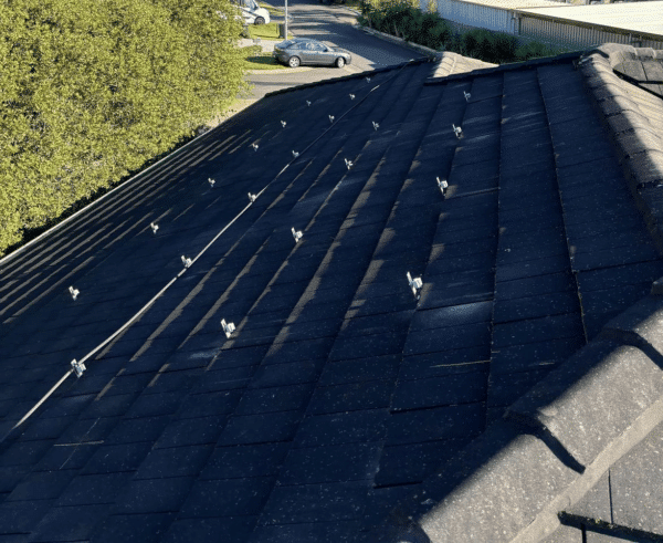 View of a tiled roof with multiple roof anchor points installed. A road, vegetation, and industrial buildings are visible in the background.