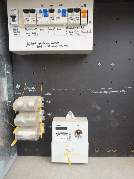 Electrical panel with labeled circuit breakers, service fuses, and a meter against a marked black surface. Notes are handwritten around the components.