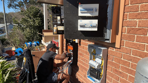 A person is working on electrical wiring outside, with various electrical panels and tools visible around them. The setting includes a brick wall and greenery.