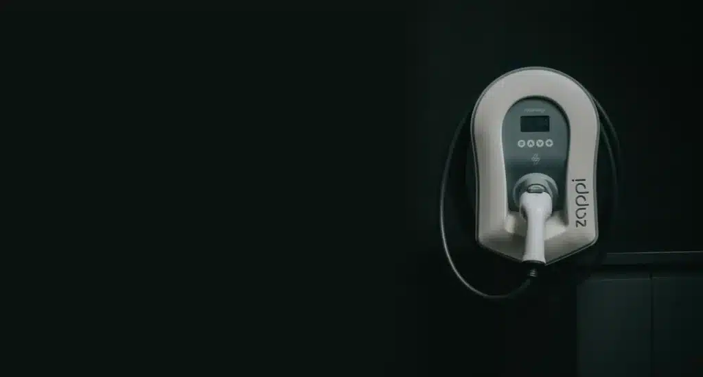 A white electric vehicle charging station with a black cord is mounted on a dark wall, with the brand name "Zaptec" clearly visible on the side of the unit.